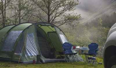 Vor einen Wald in den Bergen steht ein Zelt mit zwei Campingstühle und einen Campingtisch in Vordergrund ist noch ein stück Kotflügel eines Autos zu sehen.