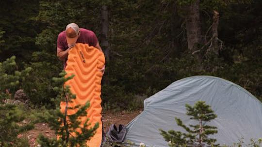 Mann pustet eine Isomatte im Wald auf. Daneben steht ein Zelt.