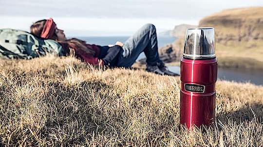 Thermosflaschen mit rastender Frau im Gras an einer Küste.